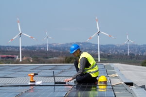 a worker in yellow vest and blue hard hat photographed installing solar panels in front of a backdrop of spinning wind turbines and mountains